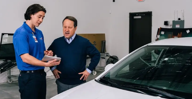 A man and male Glass Doctor service professional standing next to a white car with a cracked windshield inside an auto facility.
