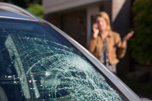 A woman stands beside a car with a shattered windshield.