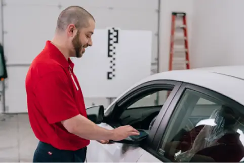 A male Glass Doctor service professional standing beside a white car by the driver’s side window.